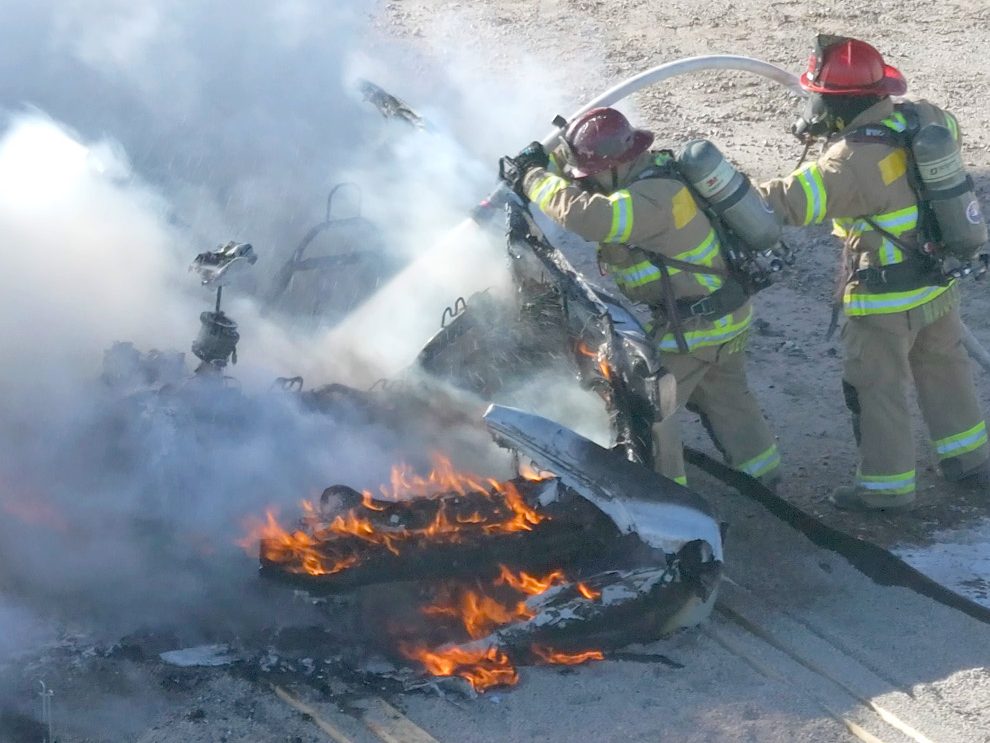 Two firefighters in full gear battling a fire that is engulfing a vehicle, with thick smoke rising into the air.